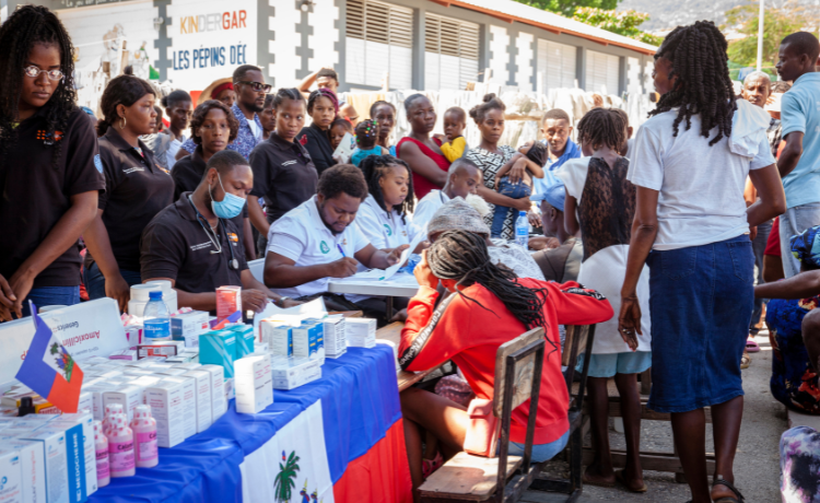 A crowd is gathered around health workers seated at a table, filling out prescriptions